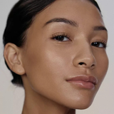 A close-up portrait of a woman with smooth, radiant medium-to-deep tan skin, dark hair pulled back, and subtle glossy lips against a soft light gray background.