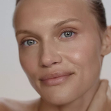 Close-up portrait of a woman with smooth, radiant skin and subtle rosy blush, looking directly at the camera against a soft light gray background.